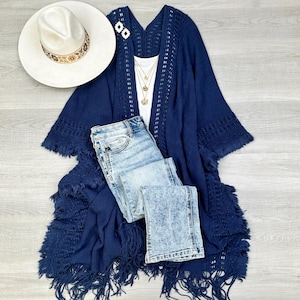 May include: A navy blue crochet fringe cardigan with a white crochet trim. The cardigan is open in the front and has a relaxed fit. It is paired with a white tank top, light blue jeans, and a gold necklace.