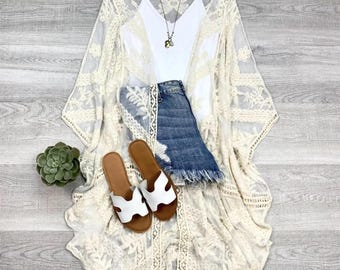 May include: A white crochet lace kimono with a white vest top, denim shorts, and white sandals.