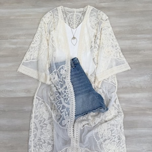 May include: A white crochet lace kimono with a delicate floral pattern. The kimono has long sleeves and a flowing silhouette. It is paired with a white tank top and denim shorts.
