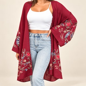 May include: A deep burgundy open-front kimono featuring wide, flared sleeves and intricate floral embroidery in shades of red, pink, blue, and green. The garment has a loose, flowing silhouette with detailed stitching along the hem and sleeves.