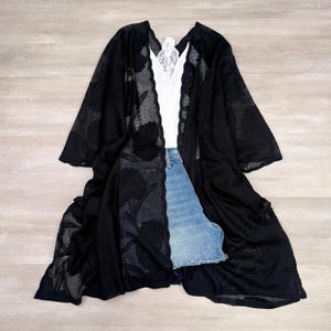 May include: A black open-front kimono featuring a sheer floral lace pattern with wide sleeves, layered over a white camisole with lace trim at the neckline and a pair of light blue denim shorts with a frayed hem and visible metal button closure.