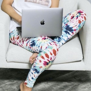 May include: A person wearing tie-dye leggings sits on a white couch and uses a silver laptop computer.