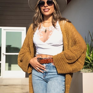 May include: A woman wearing a white crochet top, a brown cardigan, blue jeans, a brown belt, and a white fedora. She is wearing sunglasses and smiling.
