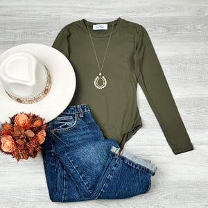 May include: A long-sleeved olive green bodysuit with a gold necklace featuring a teardrop pendant. The bodysuit is paired with blue denim jeans and a white fedora hat.