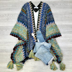 May include: A blue and green knitted poncho with a geometric pattern and fringe tassels. The poncho has a V-neck and is made of a soft, fuzzy material.