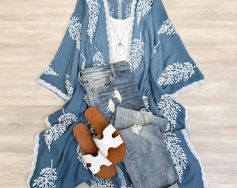 May include: A blue kimono with white floral embroidery, white lace trim, and a white tank top underneath. The kimono is paired with blue jeans and white sandals.