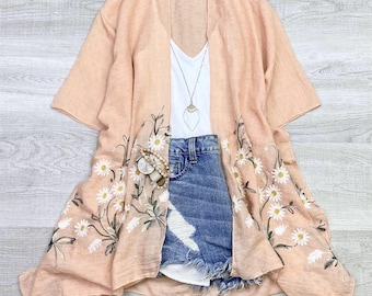 May include: A peach-coloured sheer kimono with embroidered white daisies. The kimono has a V-neck and short sleeves. It is paired with a white vest top and denim shorts.