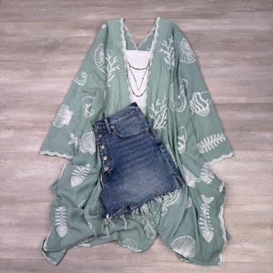 May include: A casual outfit featuring a seafoam green kimono with white seashell and coral embroidery, a white tank top, denim shorts, and a layered necklace. The shorts have a button fly and frayed hem. The kimono is draped over the other items.