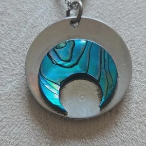 May include: A silver-toned circular pendant necklace featuring a crescent-shaped inlay of iridescent blue and green abalone shell. The pendant hangs from a matching silver-toned metal chain, showcasing a smooth metallic finish and a distinct crescent cutout.