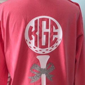 Puede incluir: Camiseta de manga larga color coral con un diseño de pelota y tee de golf blancos. La pelota de golf presenta un monograma con las letras "KGE". Debajo del tee hay un lazo plateado con purpurina. La camiseta se muestra en un maniquí.