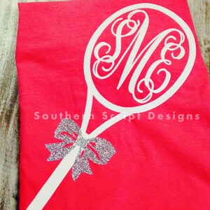 Puede incluir: Una camiseta roja brillante con un diseño de raqueta de tenis blanca, un monograma y un lazo plateado brillante. El texto "Southern Script Designs" está impreso en la parte inferior de la camiseta.