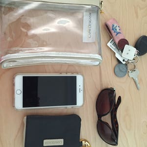 Puede incluir: Una bolsa de cosm&eacute;ticos transparente Capri Designs, un tel&eacute;fono inteligente, una cartera negra, gafas de sol y llaves est&aacute;n dispuestas sobre una superficie de madera clara. La cartera y la bolsa de cosm&eacute;ticos tienen cremalleras doradas y el logotipo de Capri Designs.