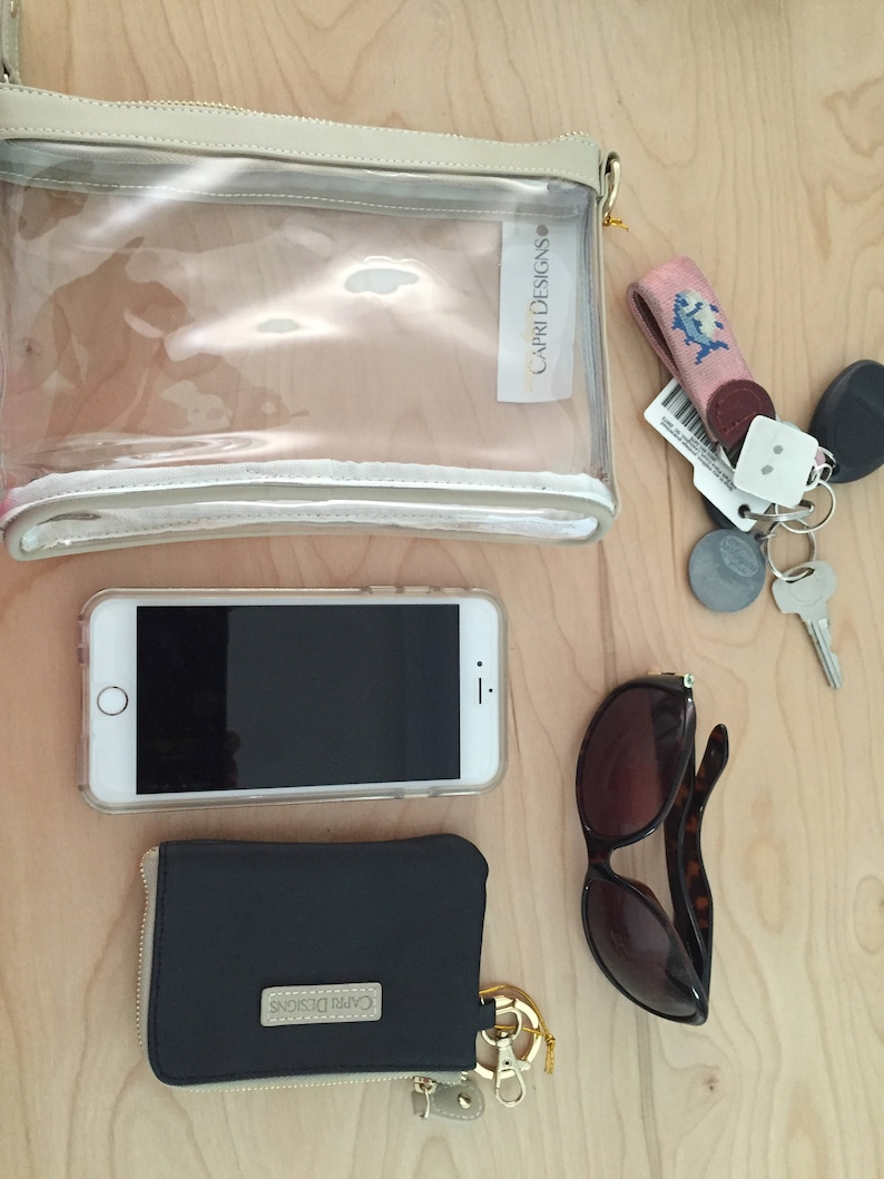 Puede incluir: Una bolsa de cosm&eacute;ticos transparente Capri Designs, un tel&eacute;fono inteligente, una cartera negra, gafas de sol y llaves est&aacute;n dispuestas sobre una superficie de madera clara. La cartera y la bolsa de cosm&eacute;ticos tienen cremalleras doradas y el logotipo de Capri Designs.