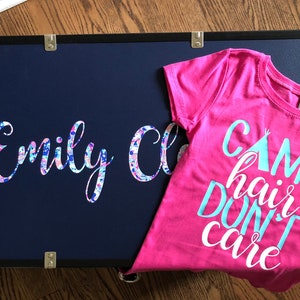 Puede incluir: Un baúl azul marino con el nombre "Emily C" en un diseño floral. Una camiseta rosa brillante con las palabras "Camp Hair Don't Care" en turquesa y blanco está al lado del baúl. El baúl tiene detalles metálicos dorados.