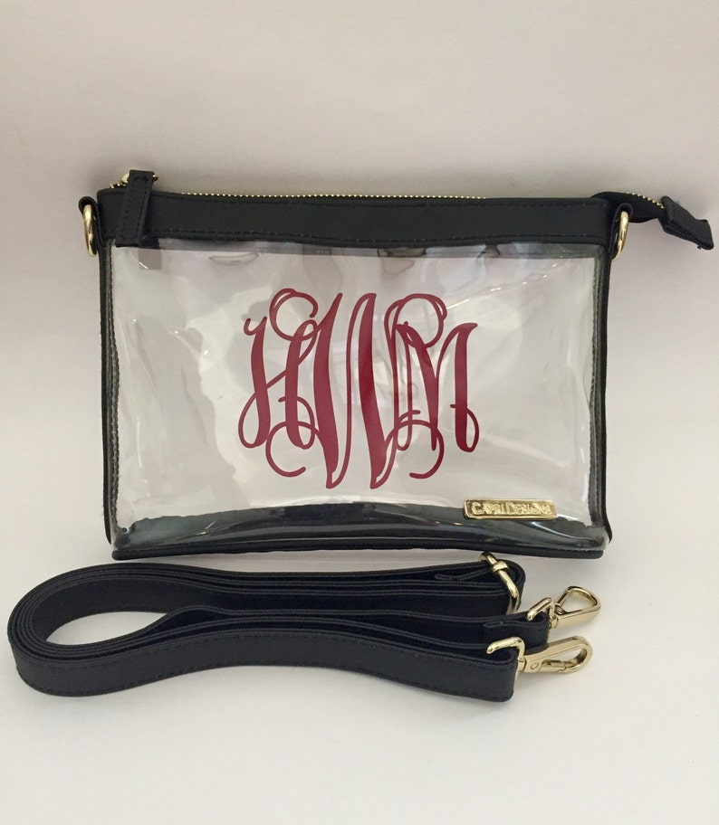 Puede incluir: Bolso cruzado transparente con parte superior y ribete negros, con un monograma burdeos. El bolso tiene cremallera y herrajes dorados, e incluye una correa para el hombro negra. Dise&ntilde;ado para llevar lo esencial.