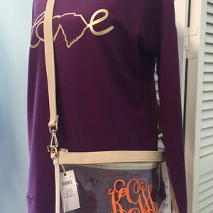 Puede incluir: Una camiseta morada de manga larga con un dise&ntilde;o dorado de "amor" y un bolso cruzado transparente. El bolso tiene un ribete color crema, un monograma naranja y una correa color crema. El bolso es de pl&aacute;stico transparente.