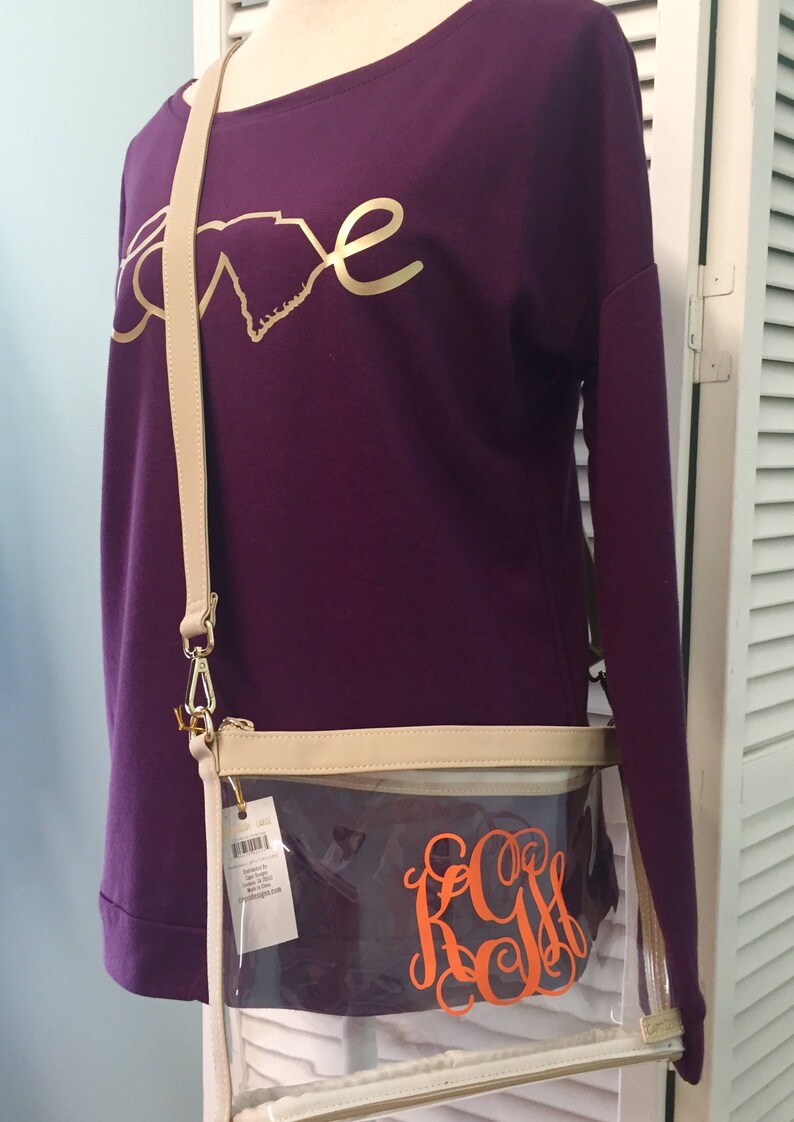 Puede incluir: Una camiseta morada de manga larga con un dise&ntilde;o dorado de "amor" y un bolso cruzado transparente. El bolso tiene un ribete color crema, un monograma naranja y una correa color crema. El bolso es de pl&aacute;stico transparente.