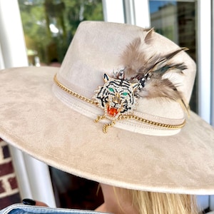 Puede incluir: Un sombrero beige de ala ancha con una banda de cadena dorada y un broche en forma de tigre. El broche está incrustado con cristales transparentes, ojos de piedra verde y una boca de piedra naranja, acentuado por plumas marrones y negras.