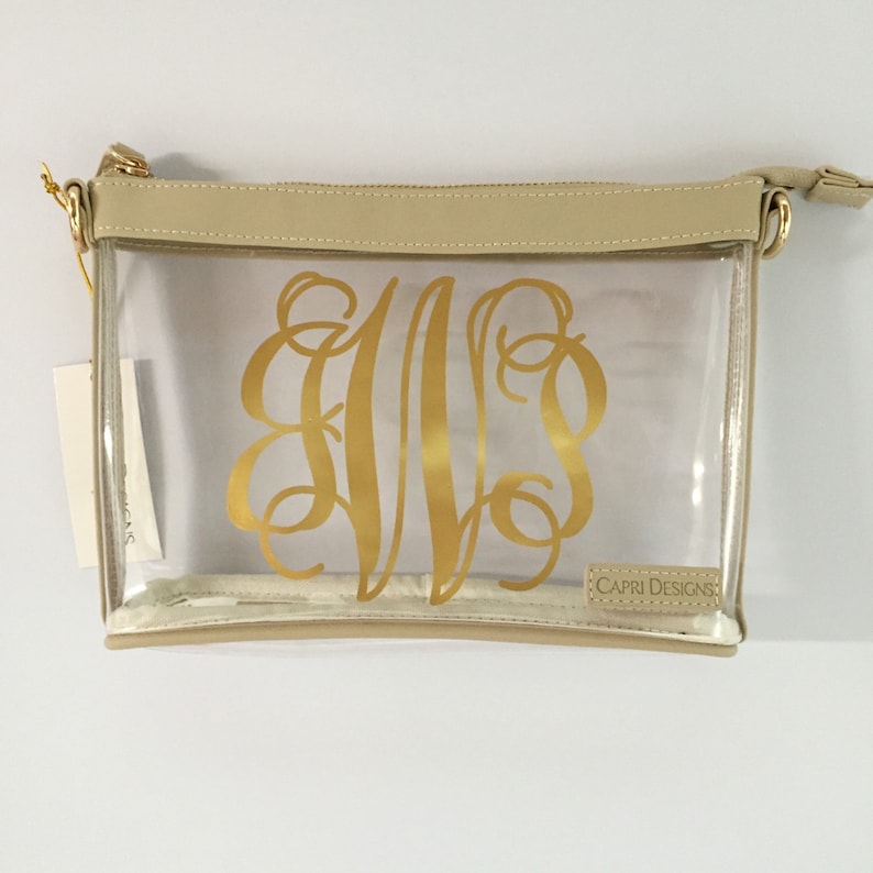 Puede incluir: Un bolso cruzado Capri Designs rectangular transparente con cremallera y correa doradas. El bolso presenta un monograma dorado y un ribete beige. Una etiqueta cuelga de la correa.