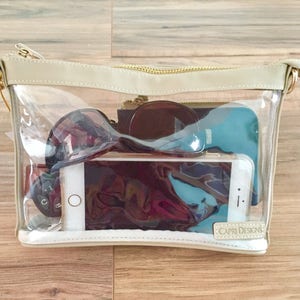 Puede incluir: Un bolso rectangular transparente de Capri Designs con ribete beige y cremallera dorada. El bolso contiene gafas de sol, un smartphone, llaves y otros objetos personales. El logotipo de Capri Designs es visible.
