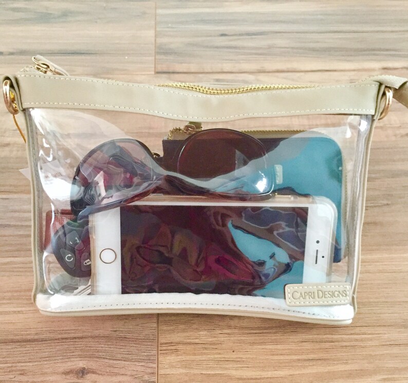 Puede incluir: Un bolso rectangular transparente de Capri Designs con ribete beige y cremallera dorada. El bolso contiene gafas de sol, un smartphone, llaves y otros objetos personales. El logotipo de Capri Designs es visible.
