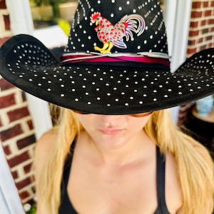 Puede incluir: Un sombrero de vaquero de fieltro negro con ala ancha y copa alta, decorado con numerosos pequeños tachuelas plateadas en la superficie. Un broche de gallo colorido con detalles en rojo, blanco y azul está prendido al frente, sobre una cinta oscura.