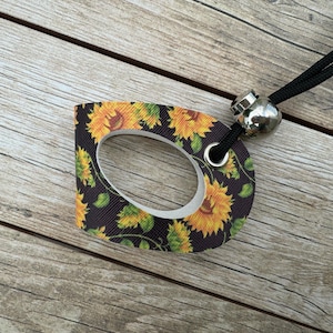 May include: A decorative tag with a sunflower pattern, featuring yellow and orange blooms with green leaves on a dark background. The tag has a white border and a metal grommet, attached to a black cord with a silver bead.