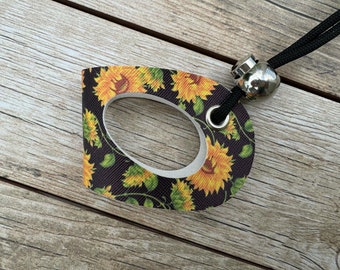 May include: A decorative tag with a sunflower pattern, featuring yellow and orange blooms with green leaves on a dark background. The tag has a white border and a metal grommet, attached to a black cord with a silver bead.
