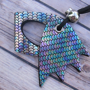 May include: A mermaid-scale patterned pendant with a black cord and silver bead. The pendant has a unique shape with a cutout design and a fin-like extension. The iridescent scales shimmer with blue, purple, and silver hues.