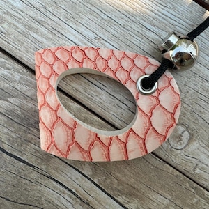 May include: A pink, scale-patterned leather tag with a central oval cutout. The tag is attached to a black cord with a silver-colored metal bead and a metal ring. The background is a wooden surface.