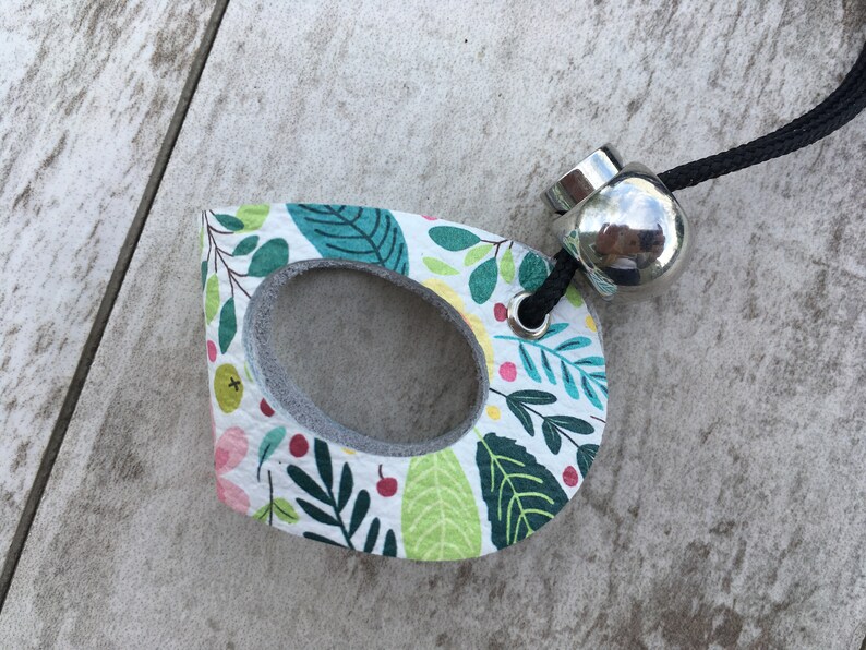 May include: A white leather charm with a floral pattern and a silver metal ring. The charm has a hole in the center and is attached to a black cord.