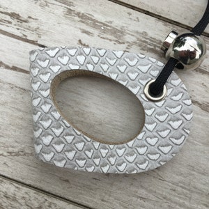 May include: A gray and white leather tag with a scale pattern and an oval cutout. The tag is attached to a black cord with a silver-colored metal bead and a metal ring.