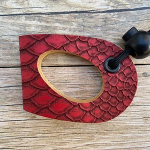 May include: A red leather-textured wooden ring with a black cord and a black bead. The ring has a hole in the center and is attached to the cord.