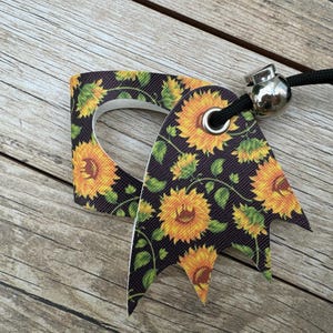 May include: A decorative tag with a sunflower pattern. The tag is black with yellow sunflowers and green leaves. It has a silver metal bead and a black cord attached.