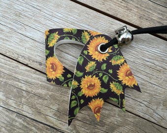 May include: A decorative tag with a sunflower pattern. The tag is black with yellow sunflowers and green leaves. It has a silver metal bead and a black cord attached.