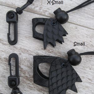 May include: Two black leather finger guards with a dragon scale design. The guards are attached to black cords with black beads and black plastic clips. The text "X-Small" and "Small" is written on the wooden surface.