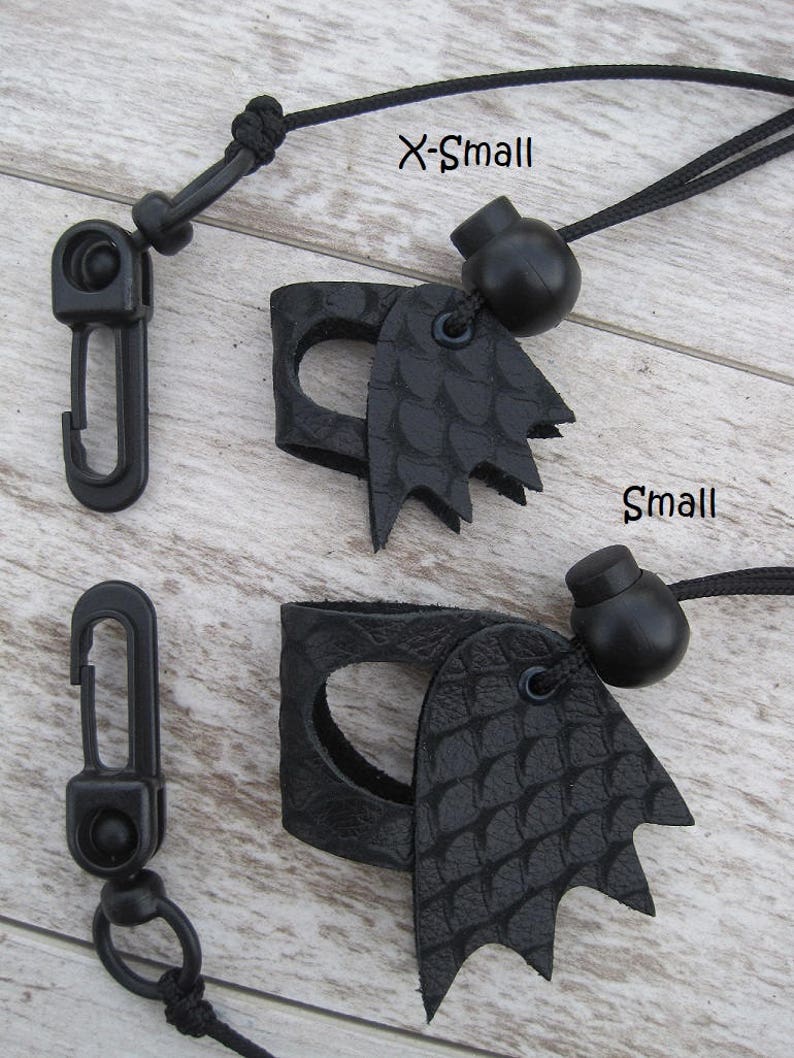 May include: Two black leather finger guards with a dragon scale design. The guards are attached to black cords with black beads and black plastic clips. The text "X-Small" and "Small" is written on the wooden surface.