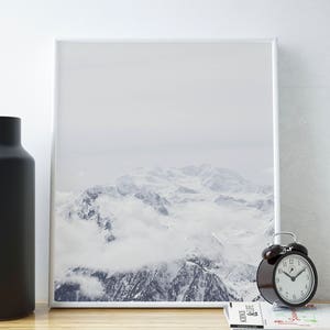 Peut inclure: Une photographie encadr&eacute;e d'une cha&icirc;ne de montagnes enneig&eacute;es avec des nuages ​​couvrant les sommets. La photo est accroch&eacute;e &agrave; un mur blanc au-dessus d'une table en bois avec un vase noir et un r&eacute;veil noir.