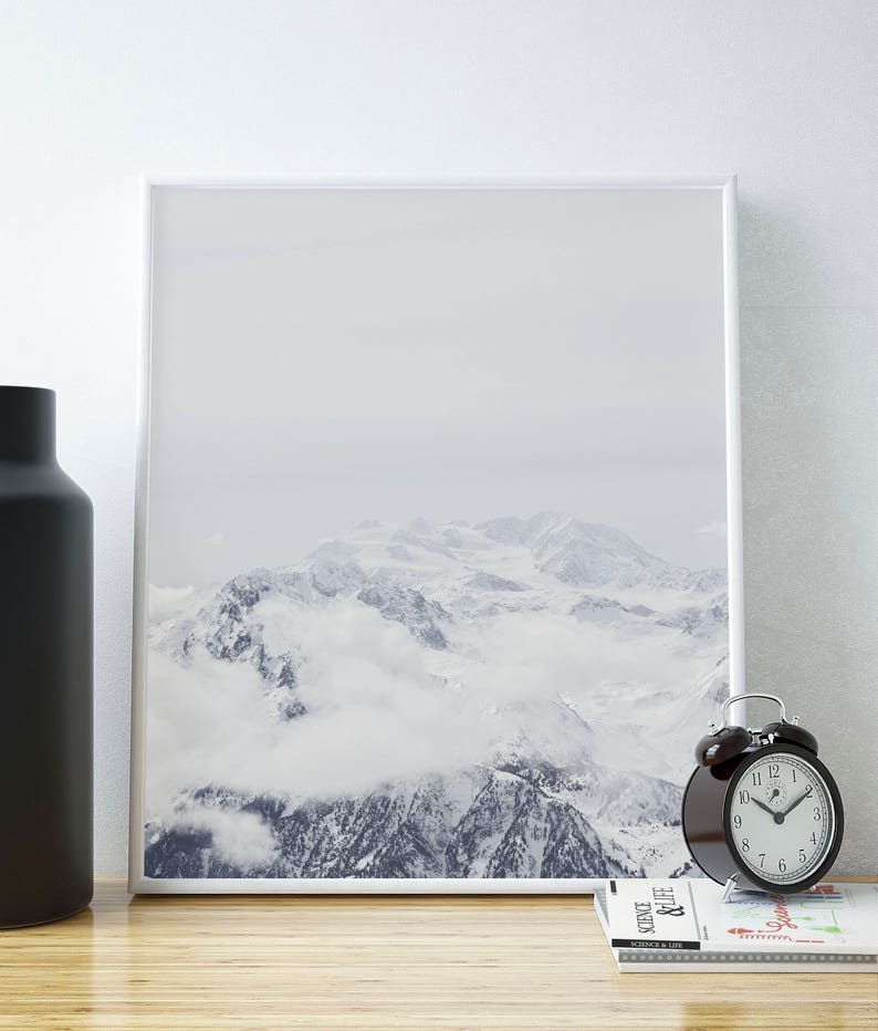 Peut inclure: Une photographie encadr&eacute;e d'une cha&icirc;ne de montagnes enneig&eacute;es avec des nuages ​​couvrant les sommets. La photo est accroch&eacute;e &agrave; un mur blanc au-dessus d'une table en bois avec un vase noir et un r&eacute;veil noir.