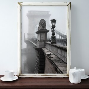 May include: A black and white photograph of a bridge in a foggy city. The bridge is made of stone and has a lion statue on top of one of the pillars. The bridge is surrounded by fog and the water below is barely visible.