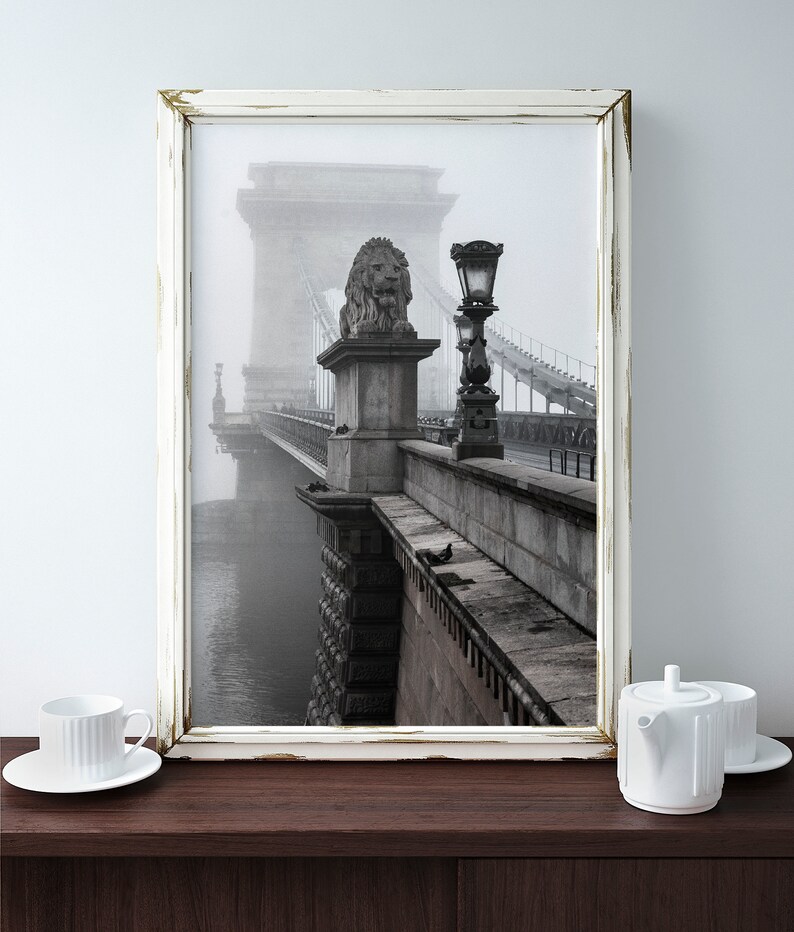 May include: A black and white photograph of a bridge in a foggy city. The bridge is made of stone and has a lion statue on top of one of the pillars. The bridge is surrounded by fog and the water below is barely visible.