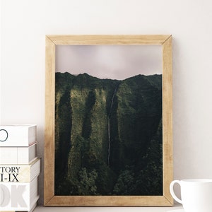 May include: A framed photograph of a lush green mountain range with a waterfall cascading down the side. The photograph is in a light brown wooden frame.