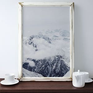 Peut inclure: Une photographie encadr&eacute;e en blanc d'une cha&icirc;ne de montagnes enneig&eacute;es avec des nuages ​​couvrant les sommets. Le cadre a une finition blanche vieillie.