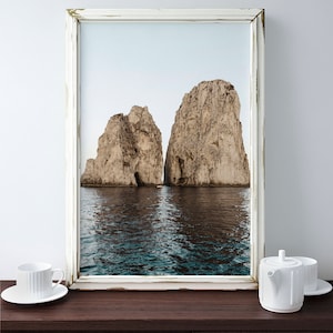 May include: A framed photograph of two large rock formations rising out of the ocean. The water is a deep blue and the sky is a light blue. The frame is white with a distressed finish.