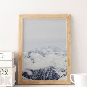 Peut inclure: Une photographie encadr&eacute;e d'une cha&icirc;ne de montagnes enneig&eacute;es avec des nuages ​​couvrant les sommets. Le cadre est en bois brun clair.