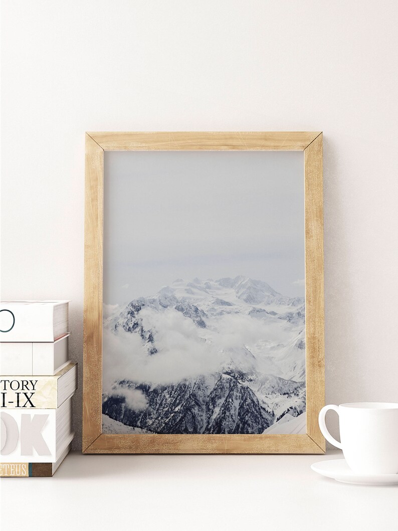 Peut inclure: Une photographie encadr&eacute;e d'une cha&icirc;ne de montagnes enneig&eacute;es avec des nuages ​​couvrant les sommets. Le cadre est en bois brun clair.