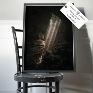Op de afbeelding: Een ingelijste print van een grotinterieur met lichtstralen die door de opening schijnen. De print zit in een zwart frame en wordt getoond op een verweerde zwarte stoel. Het label op het frame luidt "ashlee & binder PRINTABLE ART & PRINTS by ashlee binderia."