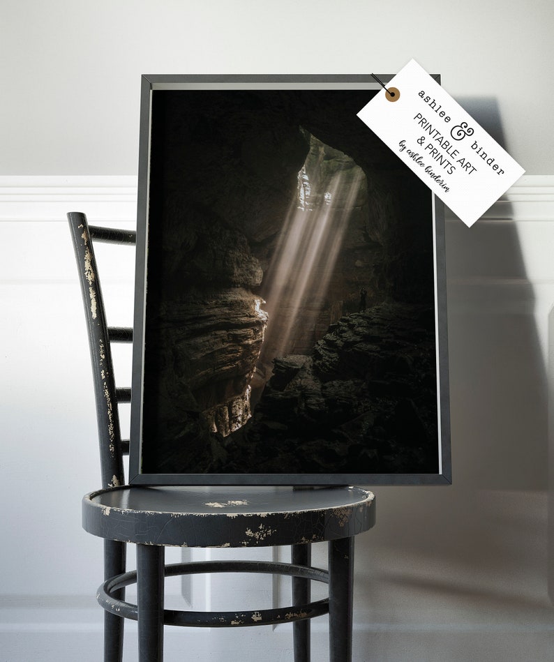 Op de afbeelding: Een ingelijste print van een grotinterieur met lichtstralen die door de opening schijnen. De print zit in een zwart frame en wordt getoond op een verweerde zwarte stoel. Het label op het frame luidt "ashlee & binder PRINTABLE ART & PRINTS by ashlee binderia."