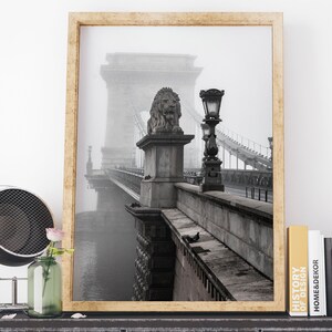 May include: A black and white photograph of a bridge with a lion statue on top of a pillar. The bridge is covered in fog and the photo is framed in a wooden frame.
