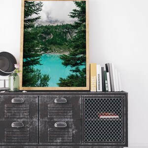 Op de afbeelding: Een ingelijste landschapsfoto van een meer omringd door bomen en bergen. Het meer is een levendige turquoise blauw en de bergen zijn bedekt met wolken. De foto hangt aan een muur boven een zwarte metalen kast met laden en een draadgaasdeur.